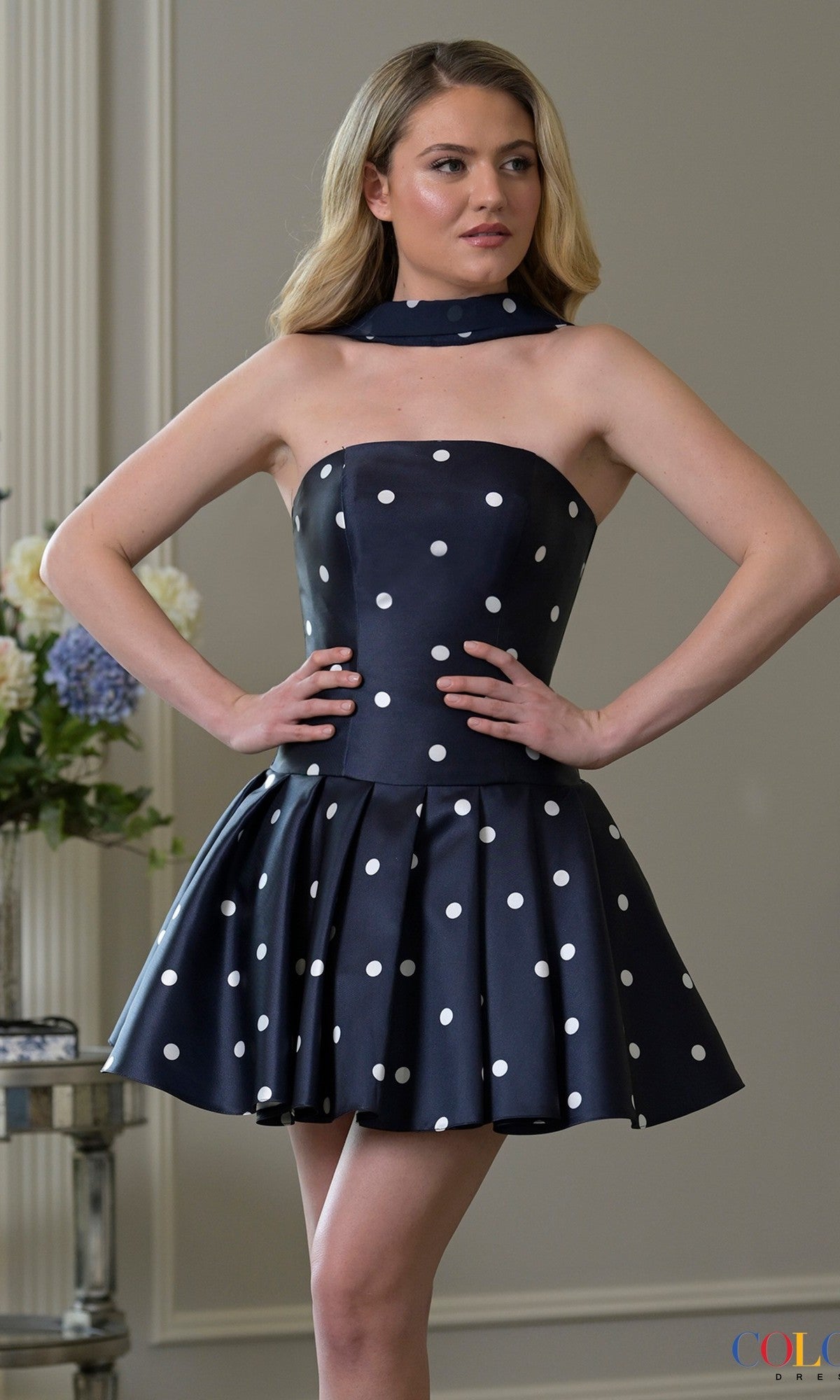 A woman stands indoors wearing the navy Short Homecoming Dress: Colors Dress 3957, featuring a strapless, polka-dot fit-and-flare silhouette and a matching neck scarf.